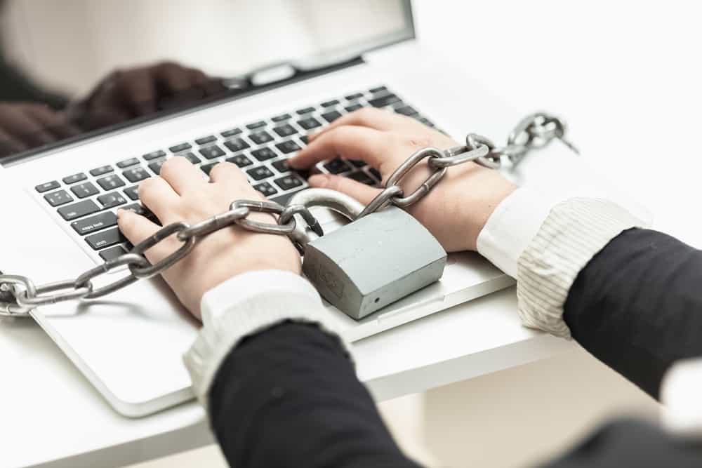 A person having his hand cuffed by a giant gray chain to a keyboard typing on a keyboard