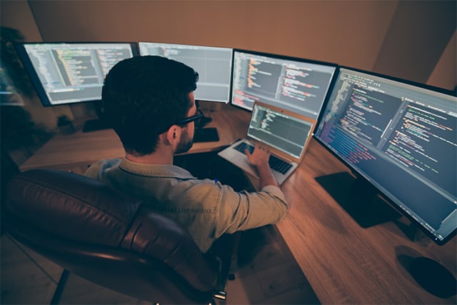 An image featuring a person using a laptop and multiple monitors while hacking