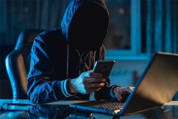 An image featuring a person wearing a hoodie and using his phone and laptop representing a cybercrime hacker concept