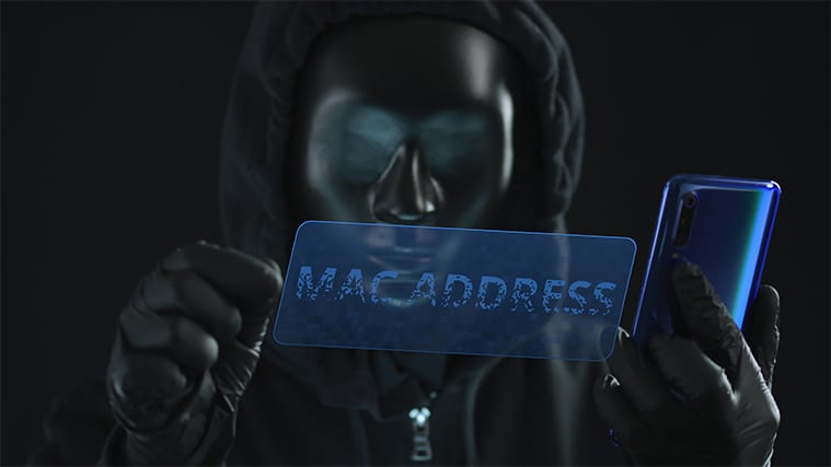An image featuring a person wearing a hoodie and a mask representing a hacker while holding a phone and has a virtual screen in front of him that says mac address